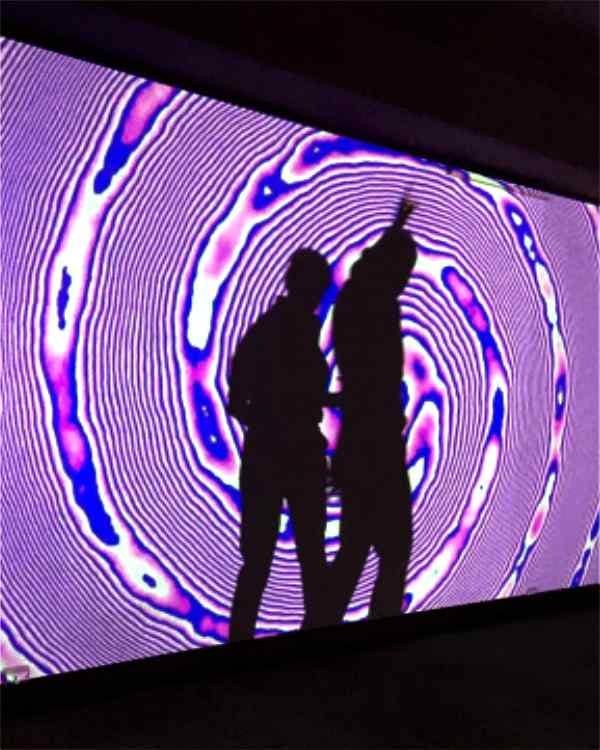 Two standing audience members casting a dancing shadow within ilithya's projection at Pop Up Art Galerie in Hamburg, Germany