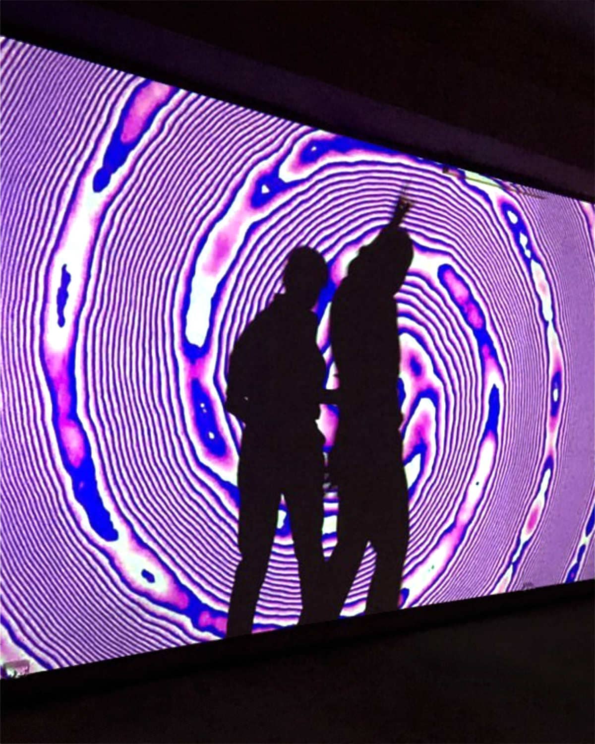 Two standing audience members casting a dancing shadow within ilithya's projection at Pop Up Art Galerie in Hamburg, Germany